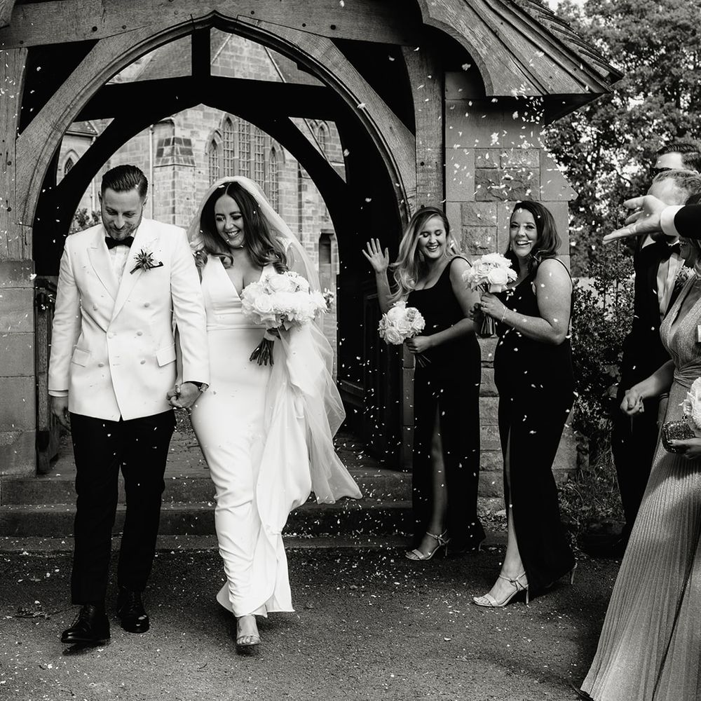 Smiling groom in white tuxedo jacket walks through confetti with smiling bride in white Made With Love wedding dress and white chapel length veil