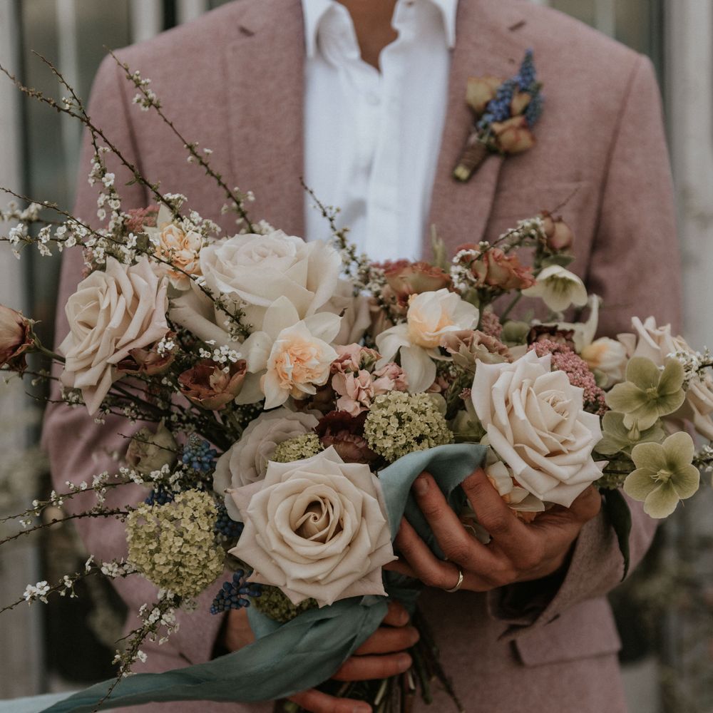 Pastel bouquet with white roses and green foliage held by groom wearing pastel pink suit 