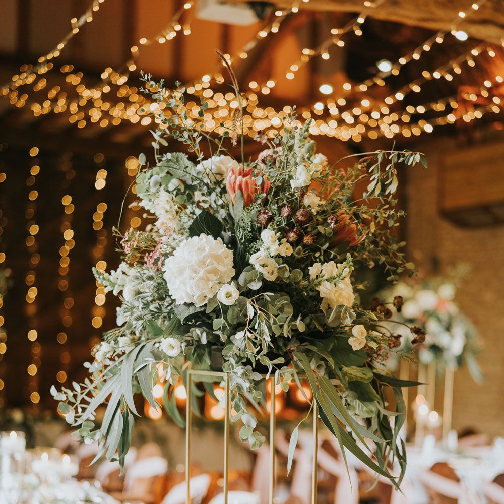 White, pink and green flower bomb on stand at long wedding breakfast table lit with strung fairy lights at Tythe Barn wedding with barn wedding flowers
