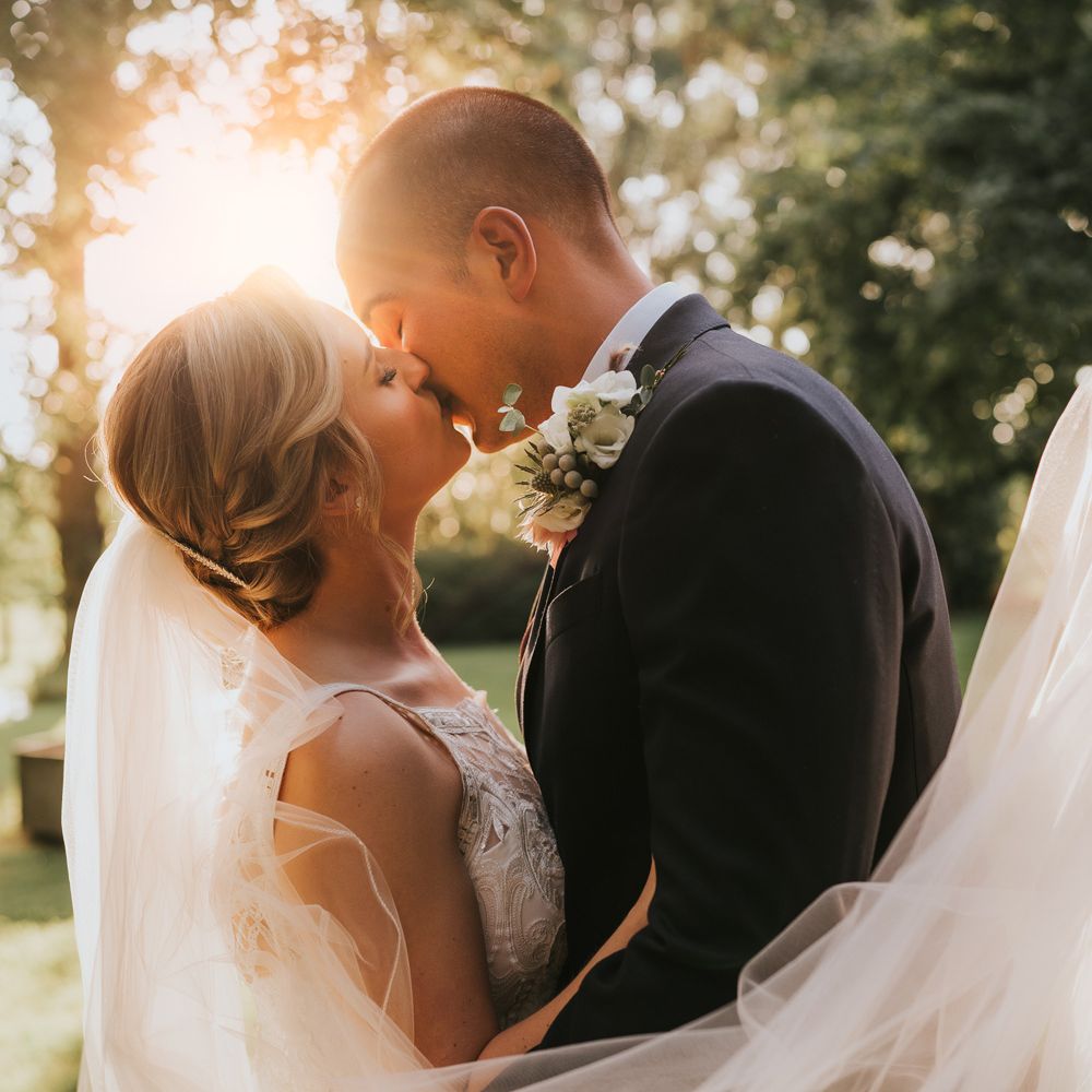 Bride in white lace Justin Alexander wedding dress and veil kisses groom in dark suit with floral buttonhole in the gardens at Tythe Barn wedding with barn wedding flowers