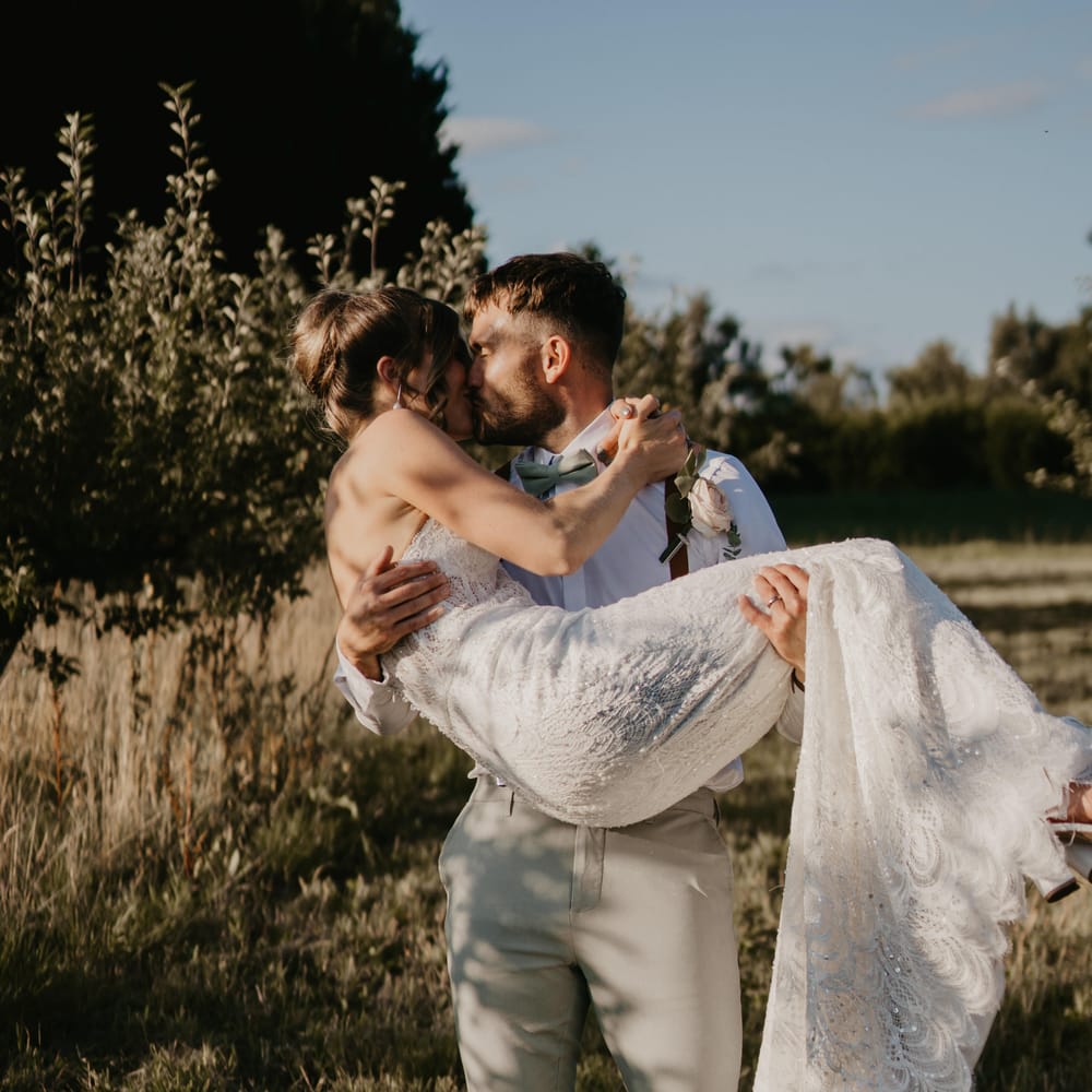 Groom carries his bride as they kiss and walk across field | Mark Bamforth Photography