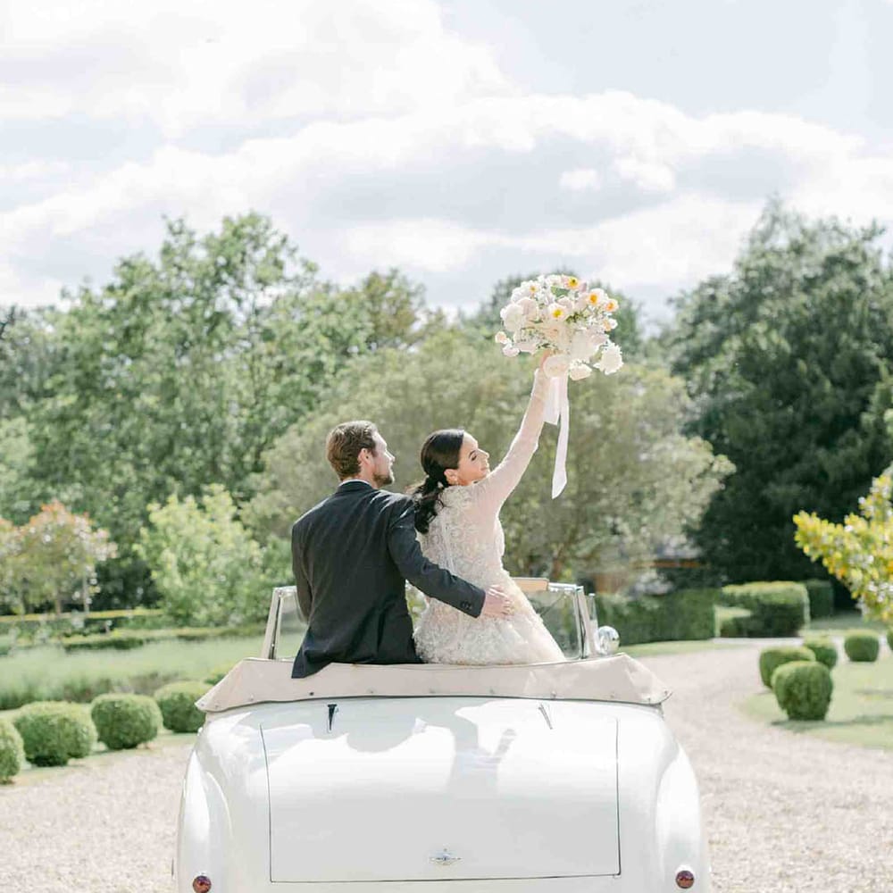 Groom embracing his bride in a long sleeve wedding dress sitting on the back of a vintage wedding car waving her bouquet in the air 