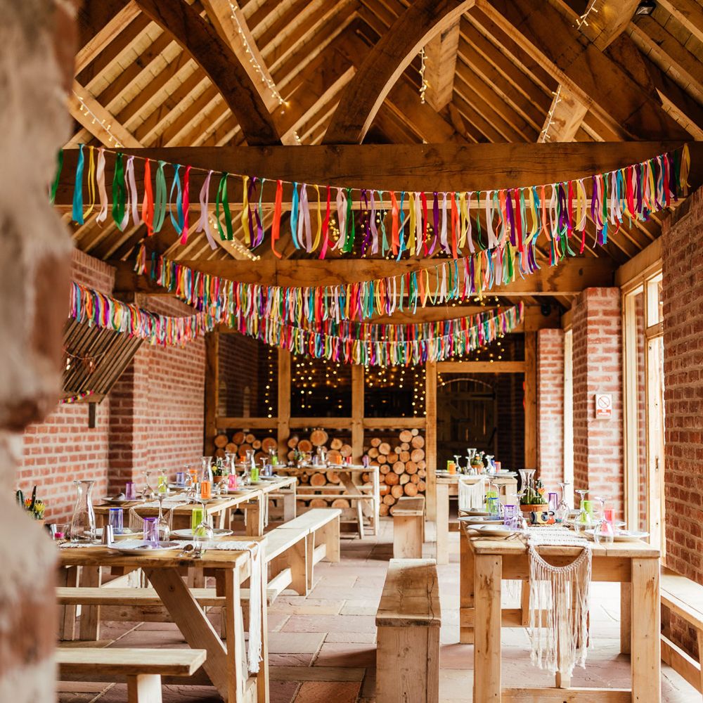 Interior of The Bridal Barn wedding venue for rustic wedding breakfast with colourful streamers, colourful glassware and wooden tables and benches