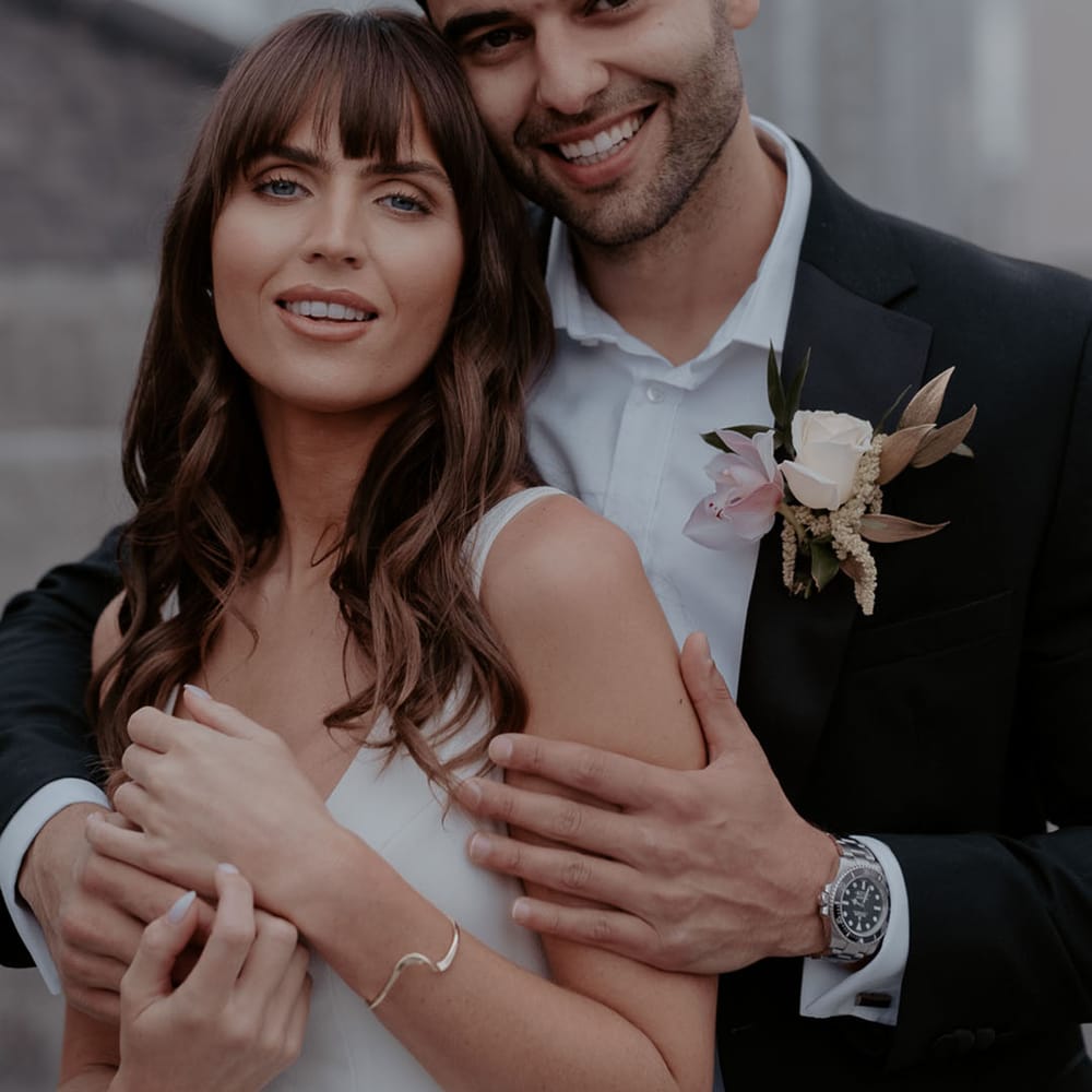 Groom in a black suit with White rose and orchid buttonhole embracing his bride with long brown wavy hair and fringe 