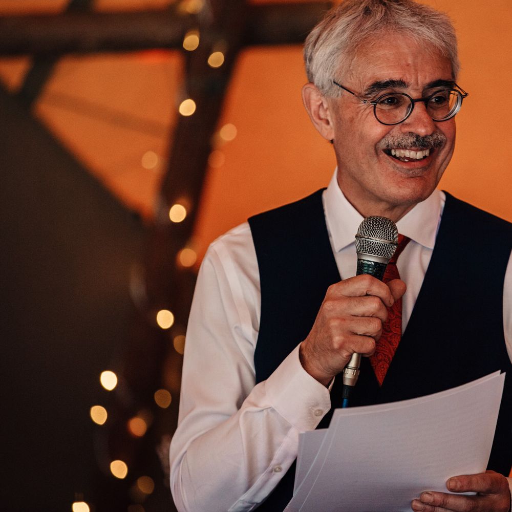 Wedding guest in white shirt, red tie and black waistcoat holds microphone as he makes speech at tipi wedding reception 