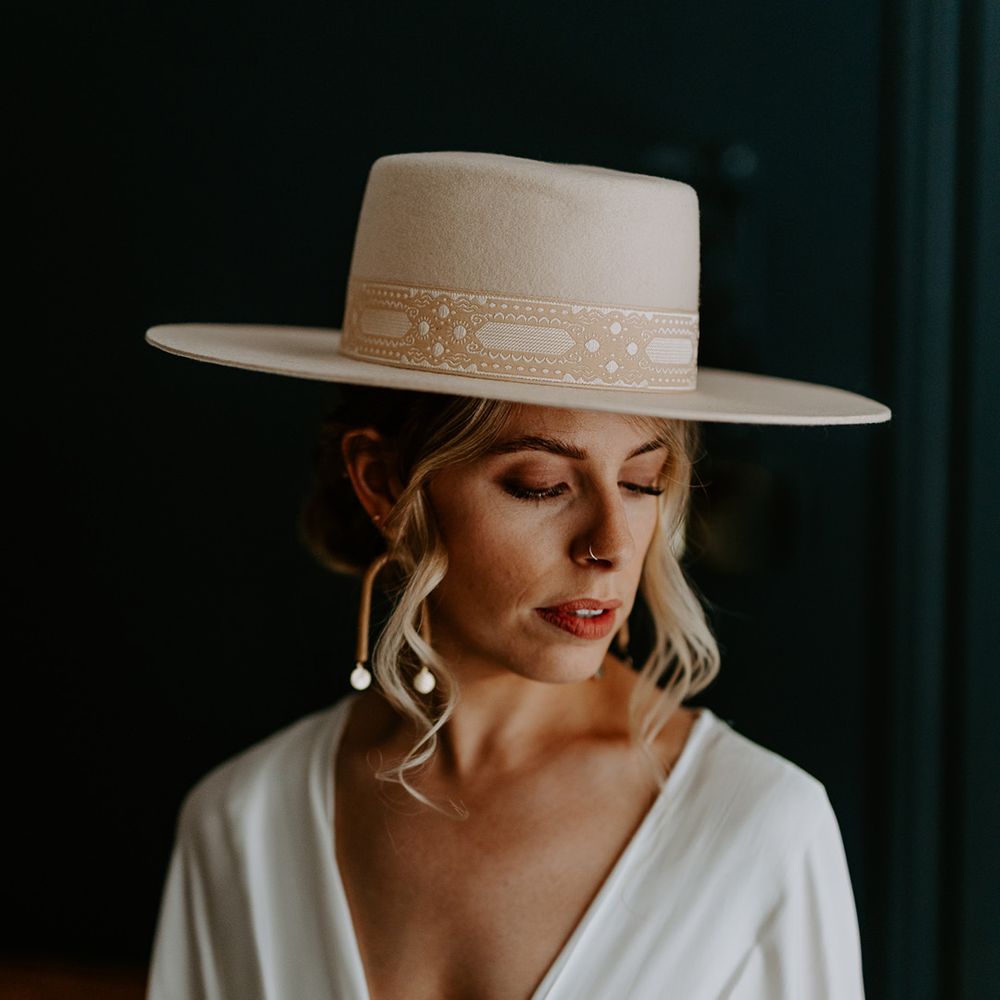 Bride wears deep V neck bridal jumpsuit with wedding hat and statement earrings