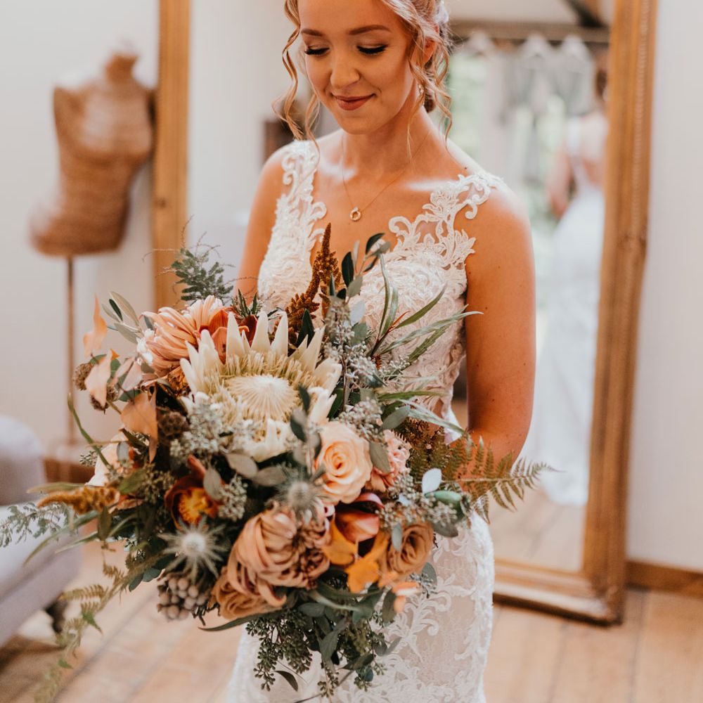 Bride with braided updo holding large mixed bridal bouquet and lace wedding dress stands in bedroom before wedding ceremony