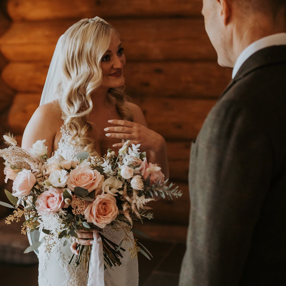 Bride with blonde hair in lace wedding dress and veil holding mixed pink and white bridal bouquet smiles at man in suit before rustic wedding