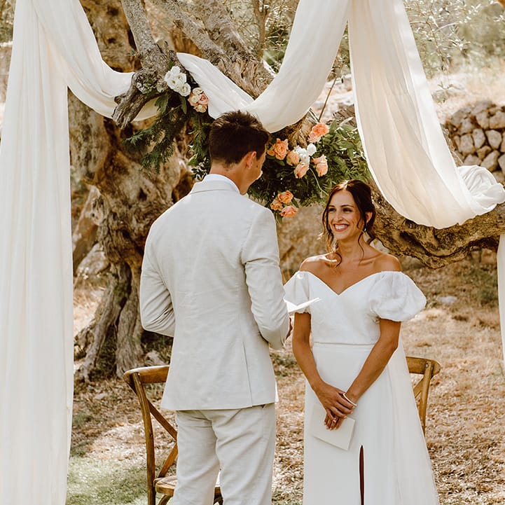Smiling bride in Alexandra Grecco wedding dress as groom in beige suit reads wedding vows