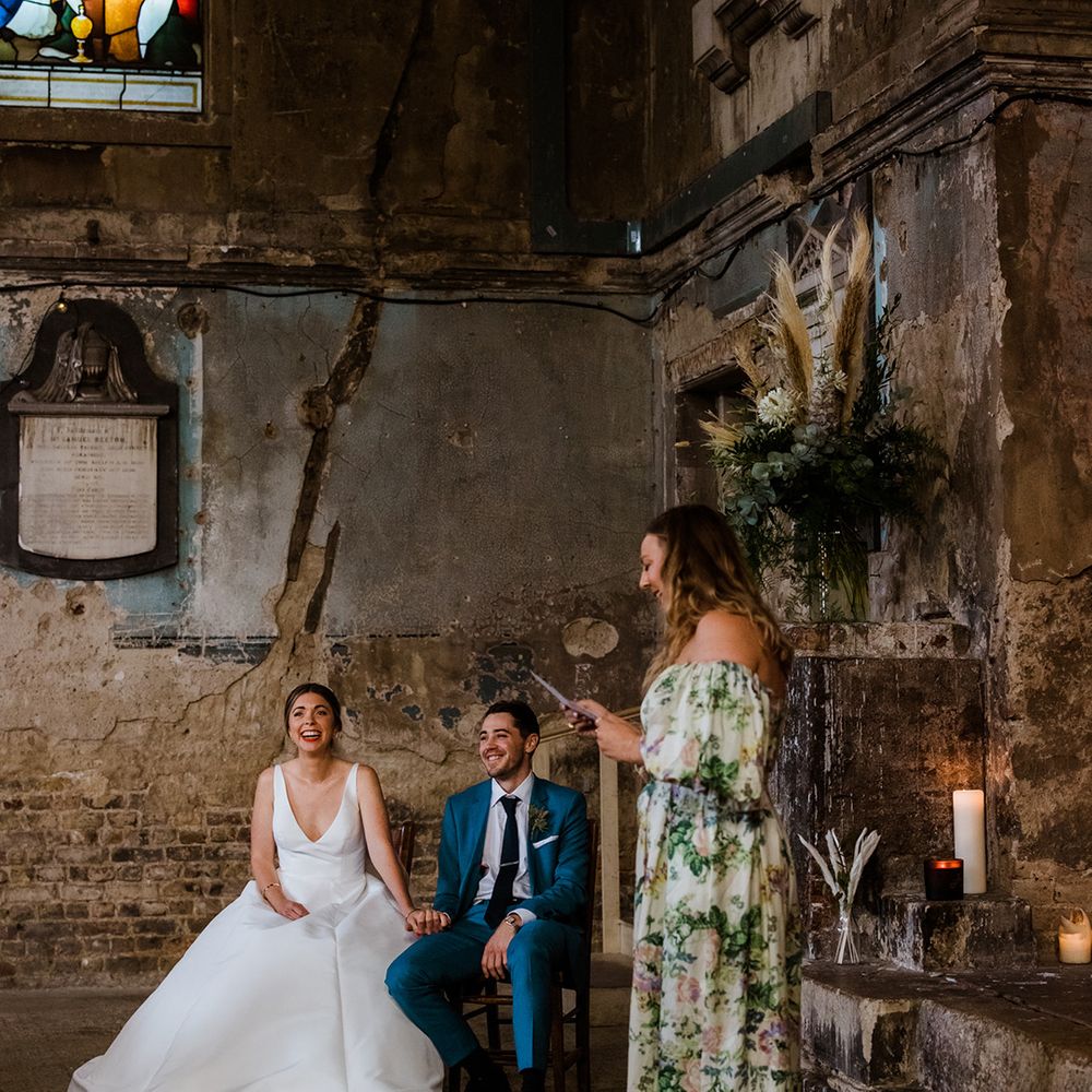 Bride in v neck wedding dress with 3D flower veil and groom in blue suit seated at industrial wedding ceremony