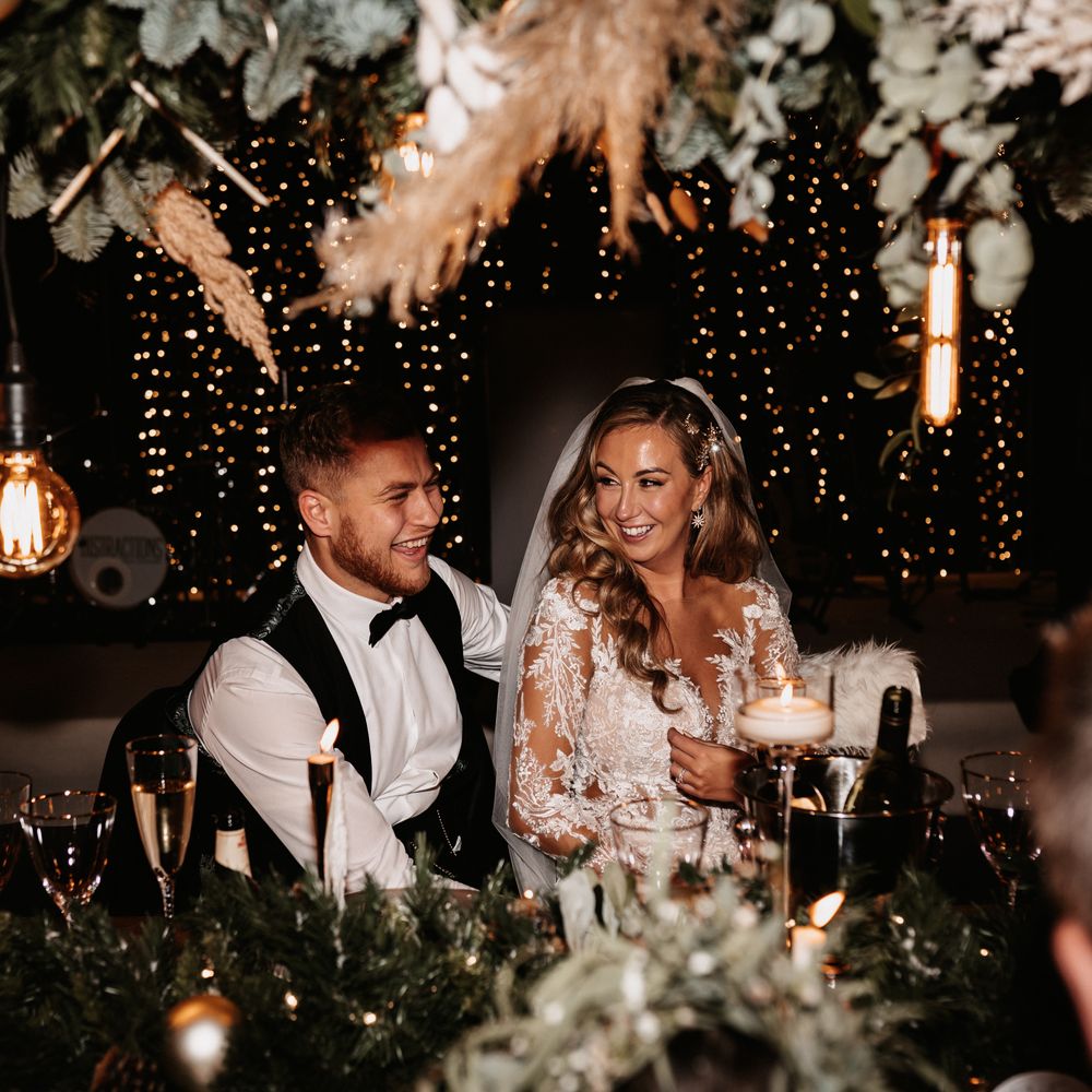 Groom in black tie and Bride seated for wedding reception with fairy lights in the background 