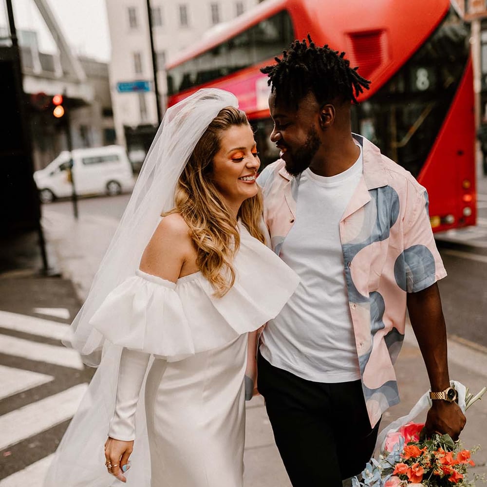 Bride in a short Bardot wedding dress with long sleeves and long veil walking through London with her groom in a patterned shirt 