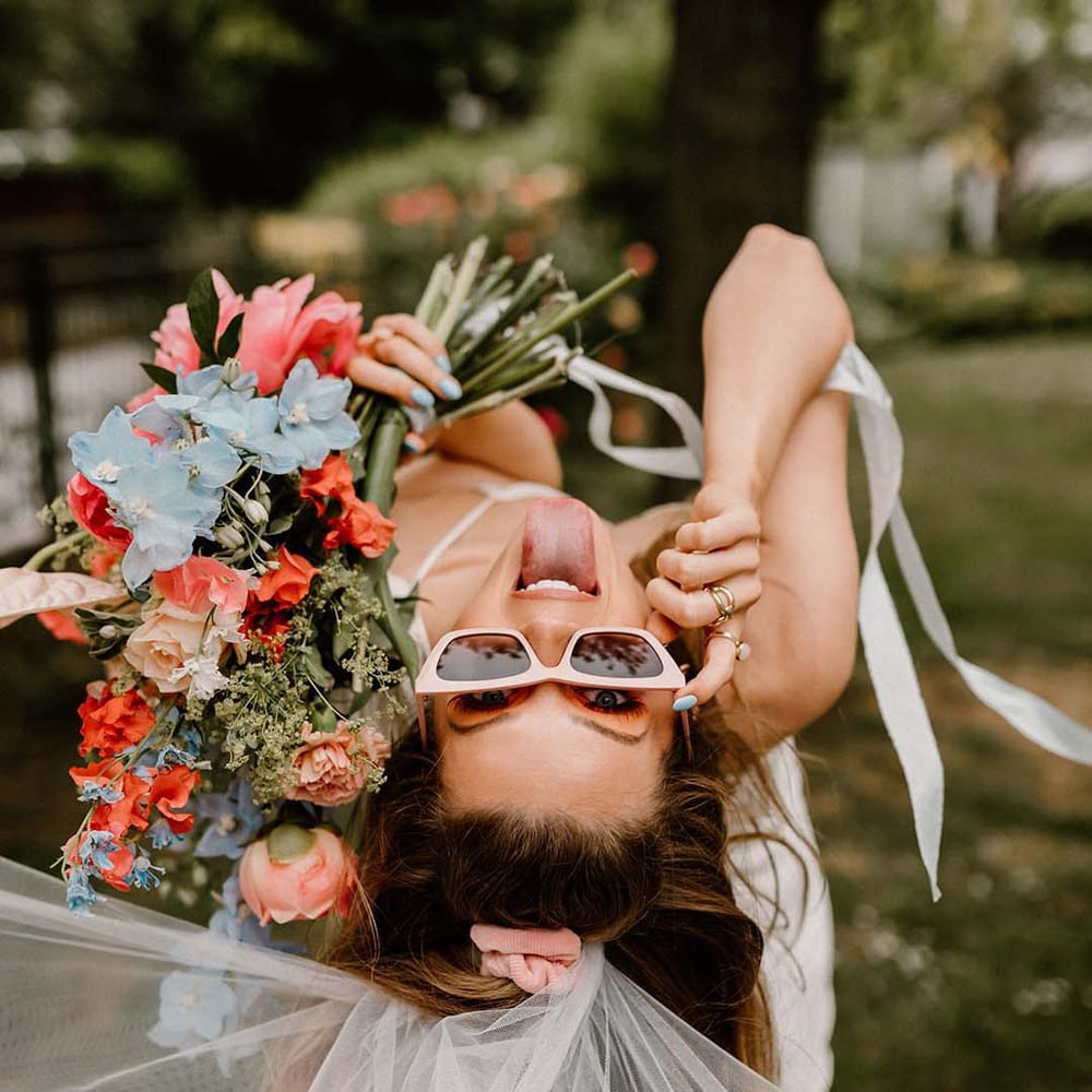 Bride leaning over in pink sunglasses holding a coral pink and blue wedding bouquet with peonies, anthuriums and delphiniums