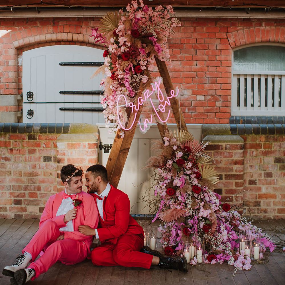 Two grooms in pink and red suits sitting in front of a wooden triangle frame altar decorated with neon sign and pink flower with dried grasses 