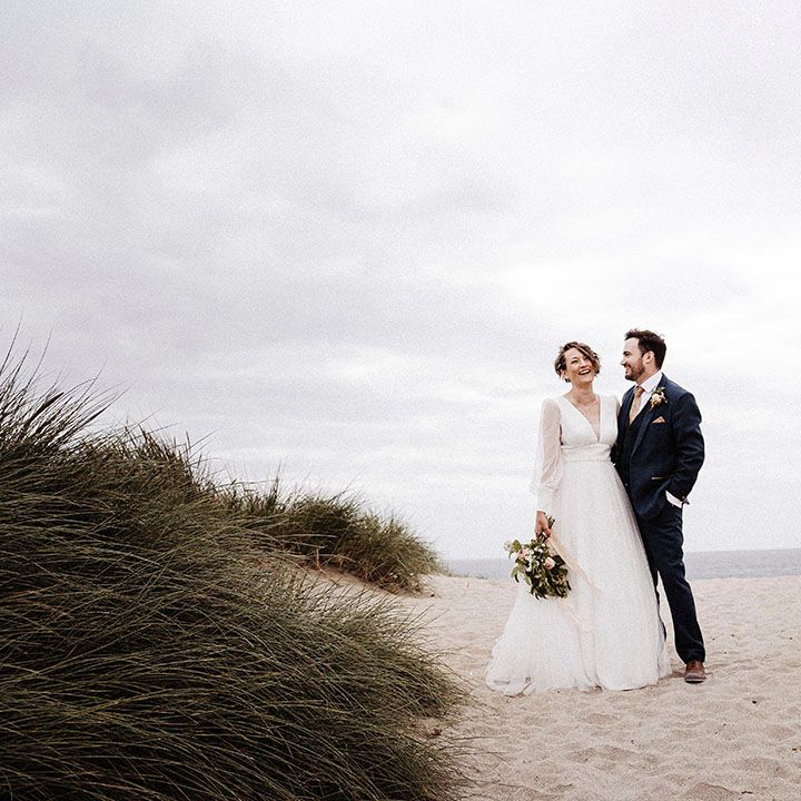 Bride and groom stand with each other on a beach in Cornwall on their wedding day with bride in tulle wedding dress and groom in blue suit