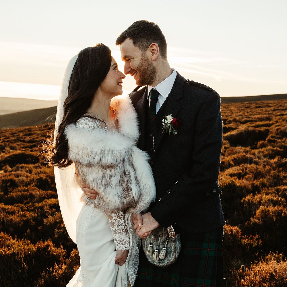 Bride wears faux fur stole and lace wedding dress stands beside her groom wearing kilt on the Scottish Highlands 