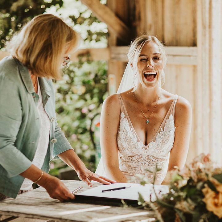 Bride gives a cheeky wink as she wears a lace Made With Love wedding dress and signs the register with the groom in a light suit 