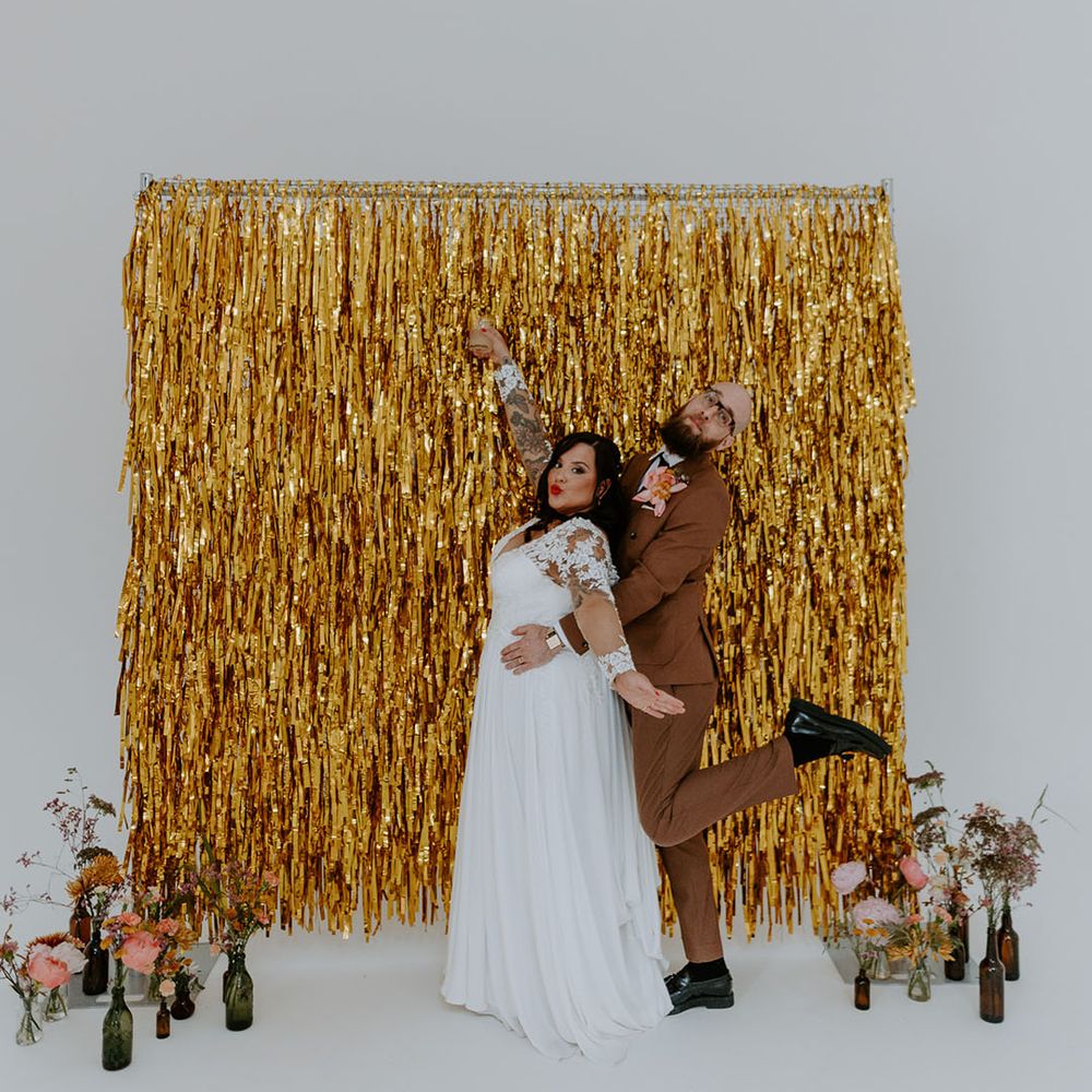Bride & groom in brown 70s styled retro suit stand in front of gold streamers wedding decor 