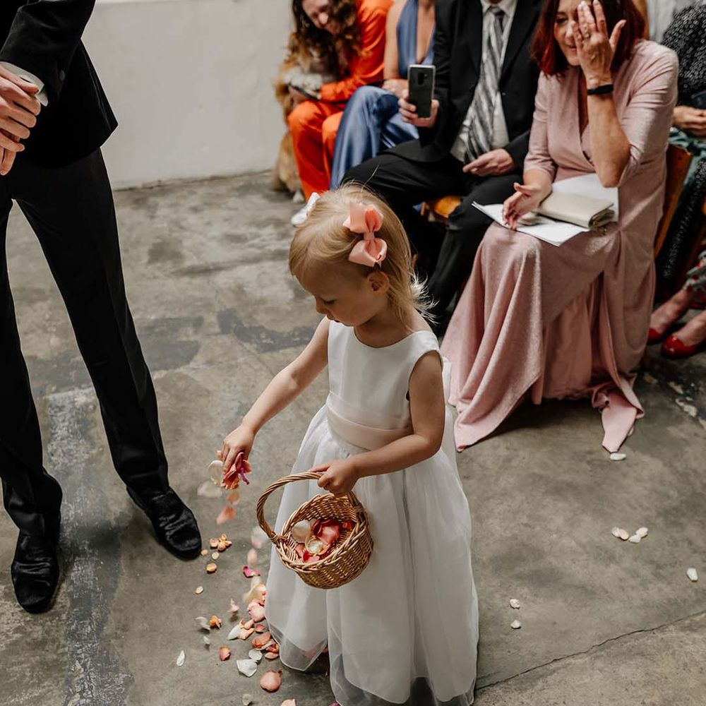 Young flower girl in white dress with light pink ribbon sprinkling flower petals at Hackney Studios