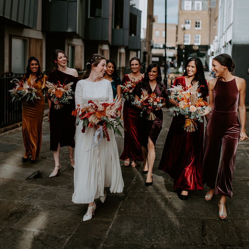 Bride in long vintage wedding dress walking with bridesmaids in mismatched velvet dresses down the streets of London, all holding large autumnal bouquets with orange, black, green and yellow roses, peonies, foliage and pampas grass