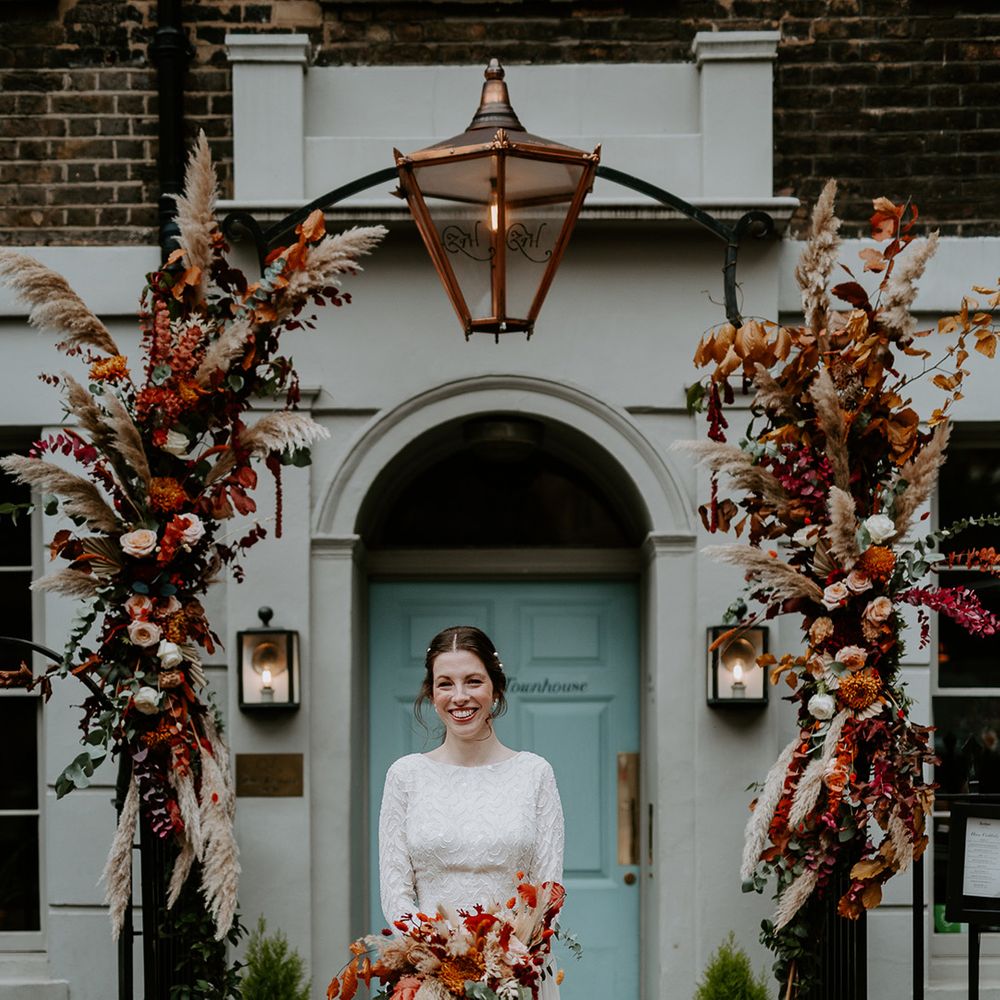 Bride in long sleeve lace vintage wedding dress and closed toe satin heels holding large autumnal bouquet and standing at The Zetter Townhouse with large floral arch with roses, peonies, foliage and pampas grass