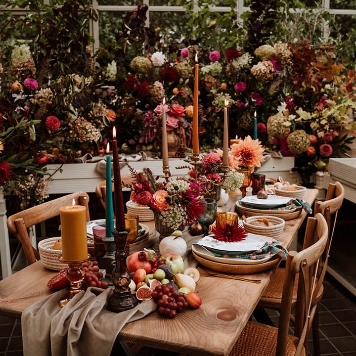 Autumnal wedding table plan with neutral toned tablerunner on rustic wooden table, orange, green and red coloured glasses, earthenware, seasonal fruit and vegetables, red, brown and green tapered candles in gold candlesticks and clear vases with autumnal coloured flowers