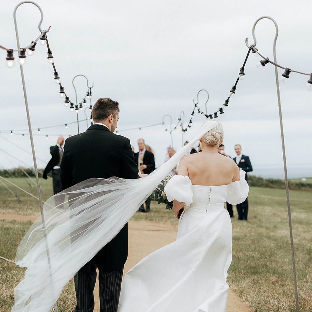 Bride wears floor length veil in her blonde hair as she walks alongside her groom toward marquee reception 