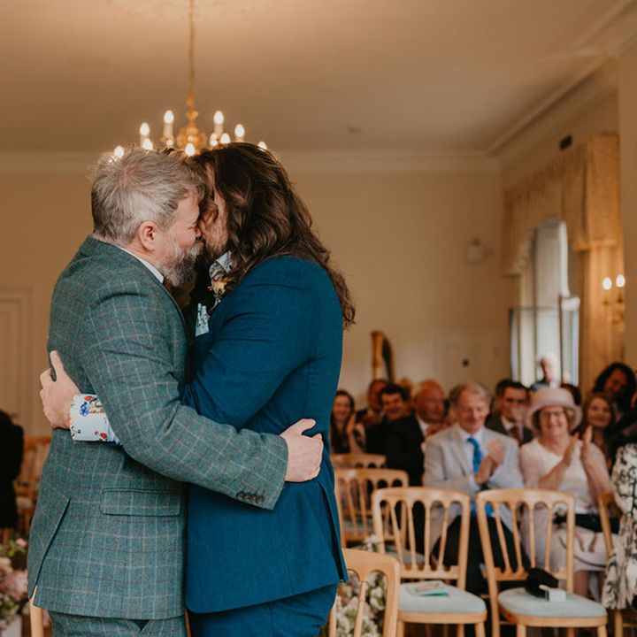 Groom in tartan suit and groom in blue suit kiss during wedding ceremony at the Cambridge Cottage in Kew Gardens 