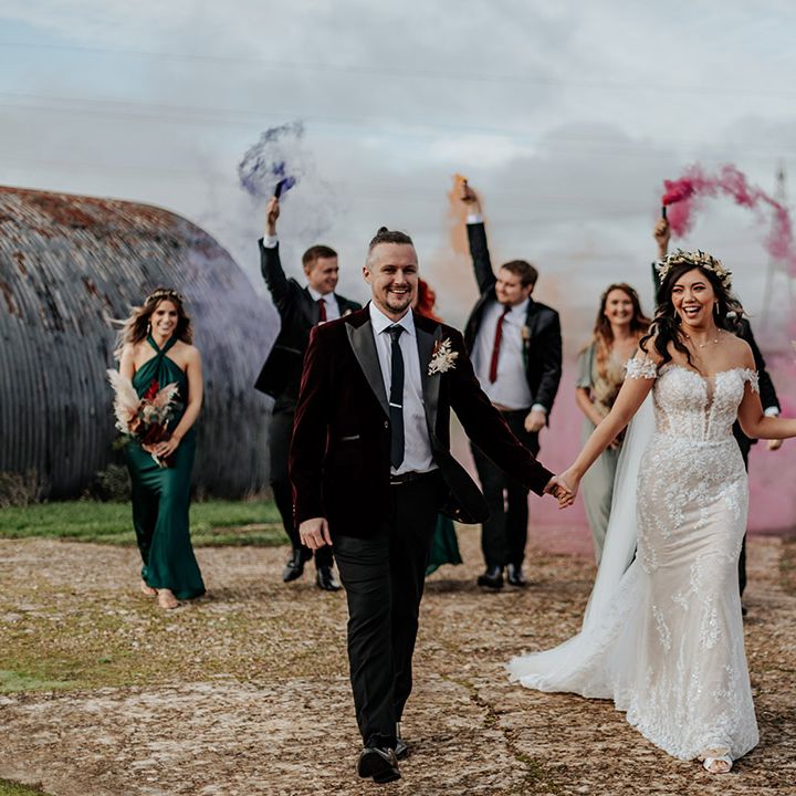 Bride in lace off the shoulder wedding dress walking with the groom in a burgundy red velvet suit jacket as smoke bombs explode 