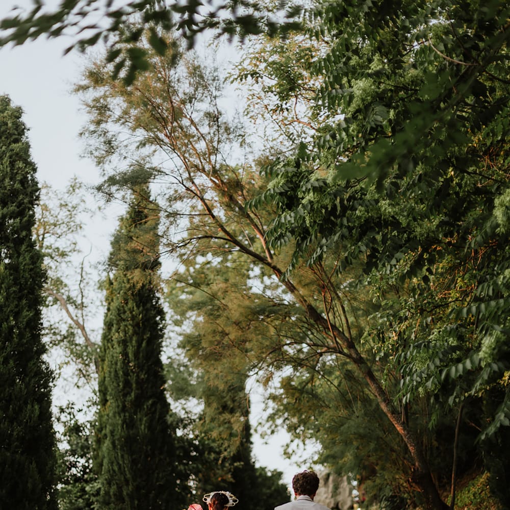 Bride and groom walk through greenery at italian destination wedding wearing white lace wedding dress, white floral structured bridal crown and greige suit
