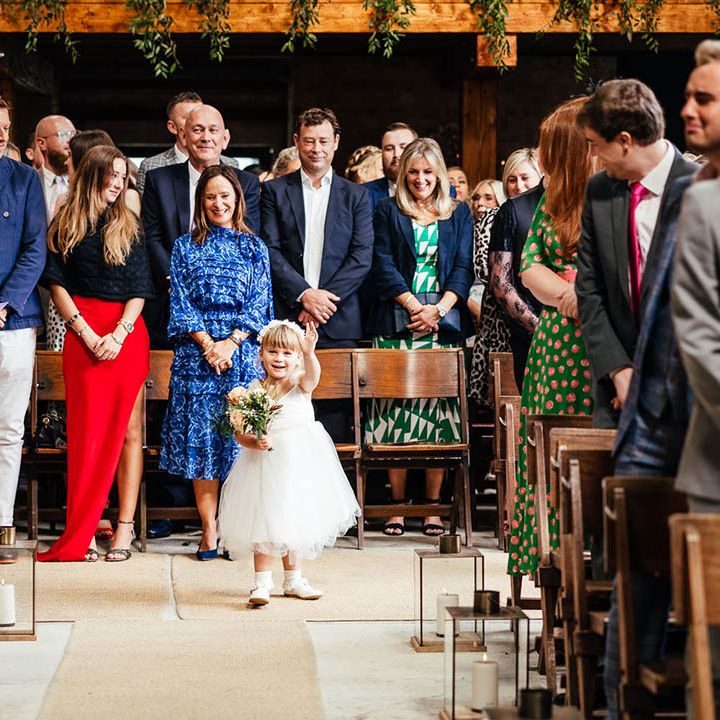 Flower girl in white tulle dress and headband walks down the aisle holding a small bouquet 