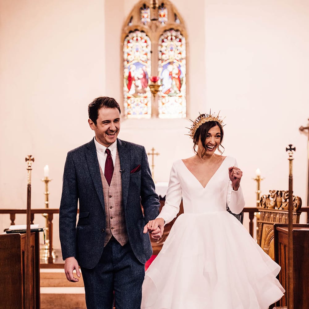 Groom in three piece navy suit walking back down the aisle of the church with the bride in ruffle wedding dress 