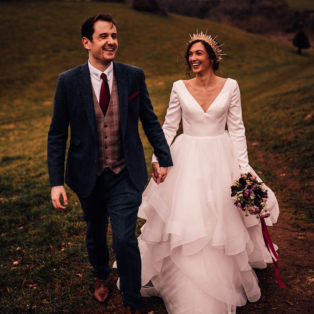 Bride in long sleeve Morilee wedding dress and groom in navy three piece suit walking hand in hand for wedding 