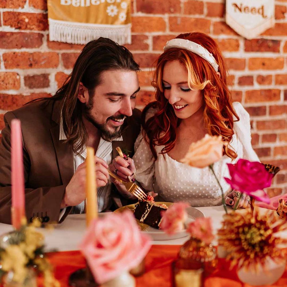 Groom in brown grooms blazer and white shirt eating wedding cake with bride in three quarter length puff sheer sleeve wedding dress with delicate polka dot overlay and square neck and white 3D applique polka dot headband at retro wedding tablescape with pink tapered candles, orange table runner and mismatched single flower vases