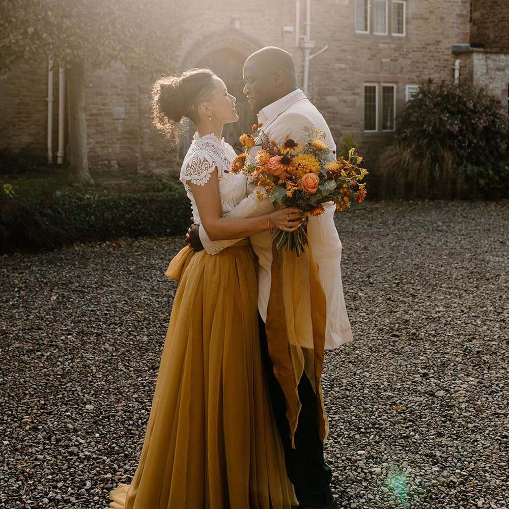 Bride in white mesh lace bridal crop top and yellow wedding skirt holding dried autumnal wedding bouquet with sunflowers, garden roses, pampas grass, eucalyptus, yellow tall kangaroo paws, orange osmanthus flowers, foliage and dried flowers embracing groom in cream grooms morning suit outside of Broadfield Court wedding venue