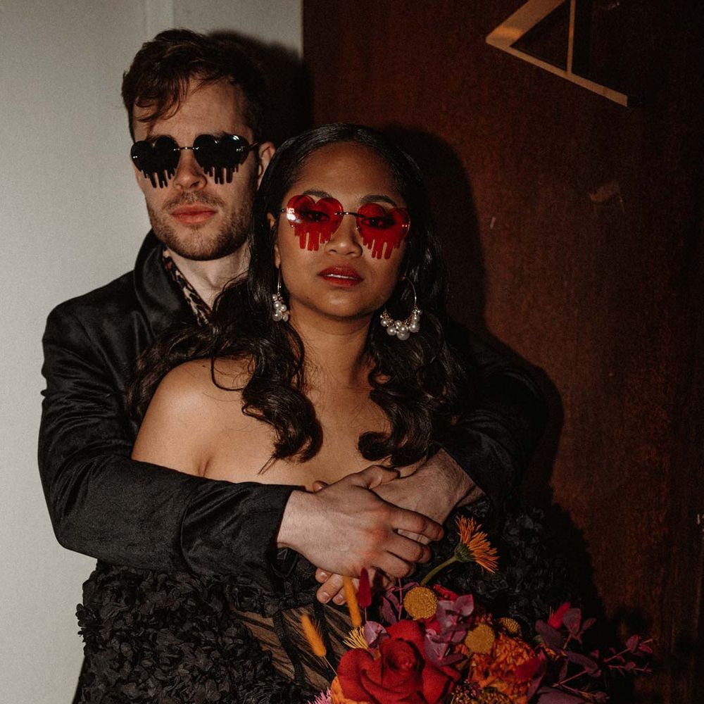 Bride in black ruffle off shoulder reception dress and red drip effect sunglasses standing with groom in black shirt and black drip effect sunglasses at German shoot with vinyl wedding arch