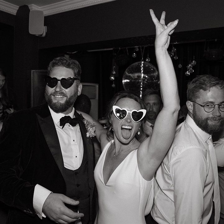 Bride wearing white heart retro wedding sunglasses dancing with the groom in a black tuxedo at the wedding reception 