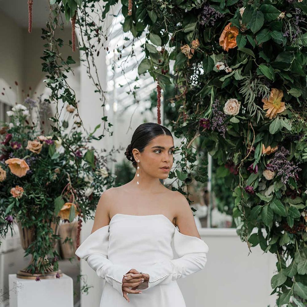 Bride in long sleeve off the shoulder wedding dress with pearl button detailing and a side slit surrounded by suspended wildflower and foliage arrangements in large gold vases at Buxted Park