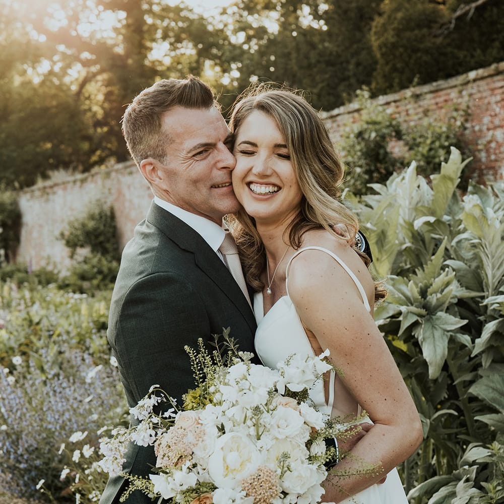 Groom in black suit jacket embracing the bride holding pale pink and white bouquet 