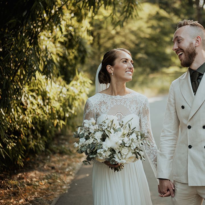 Bride in a Catherine Deane wedding dress with lace long sleeves holding hands with her groom in a beige wedding suite 