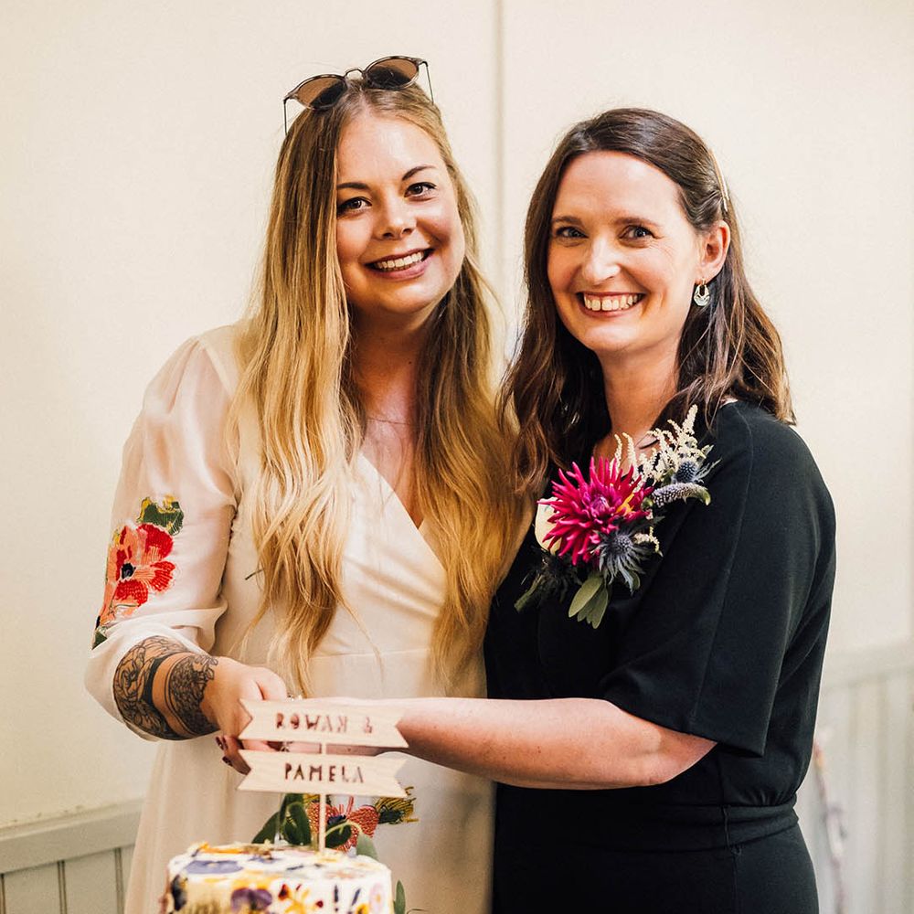Bride in colourful embroidered wedding dress and bride in black bridal jumpsuit cut their single tier wildflower pressed wedding cake 