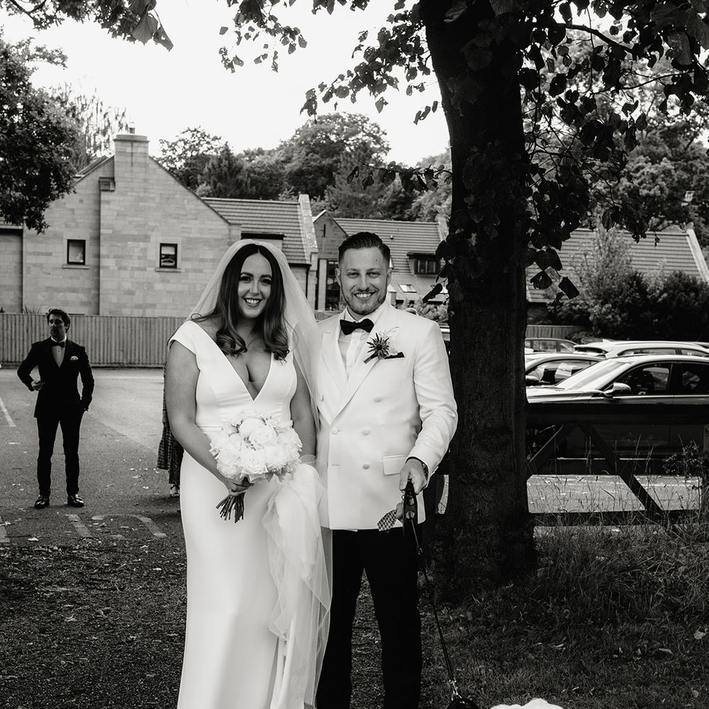 Smiling groom in white tuxedo jacket stands with smiling bride in white Made With Love wedding dress and white chapel length veil holding English bulldog on a lead