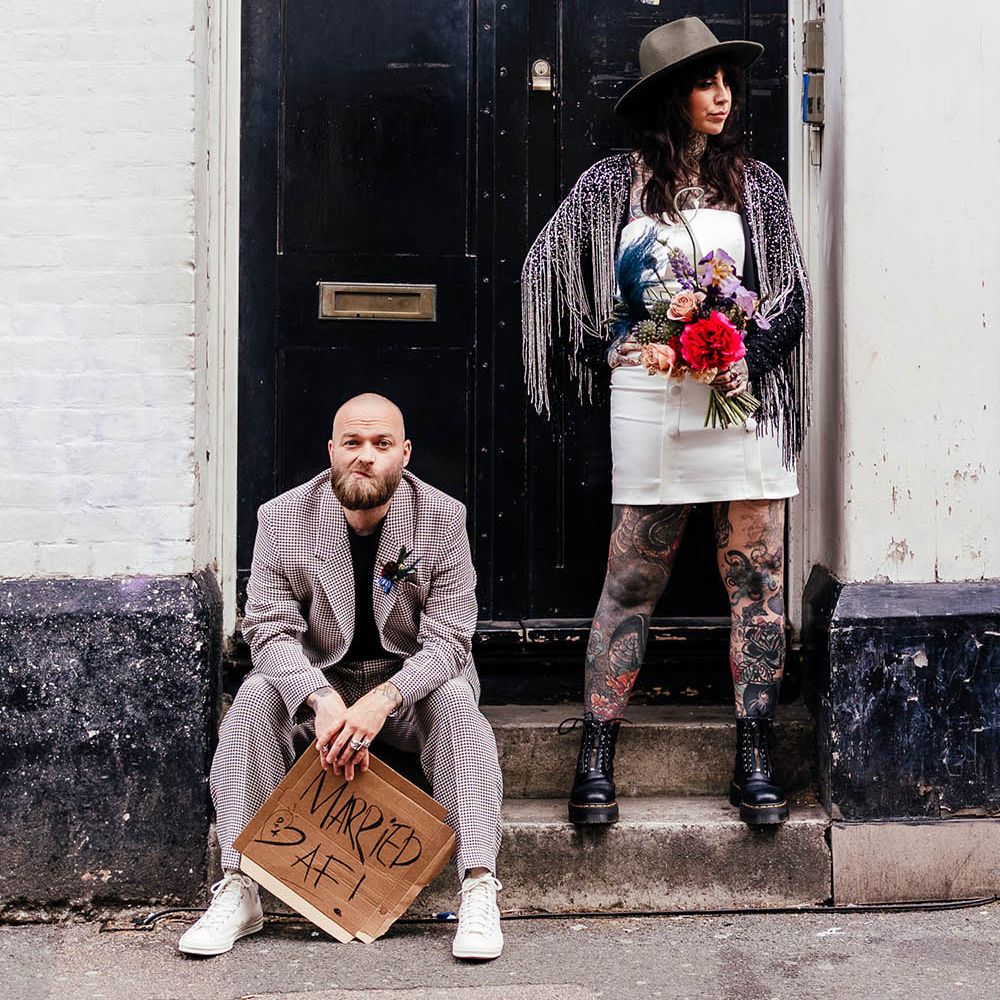 Alternative bride and groom standing in a doorway with groom in a check suit and bride in tassel jacket and hat 