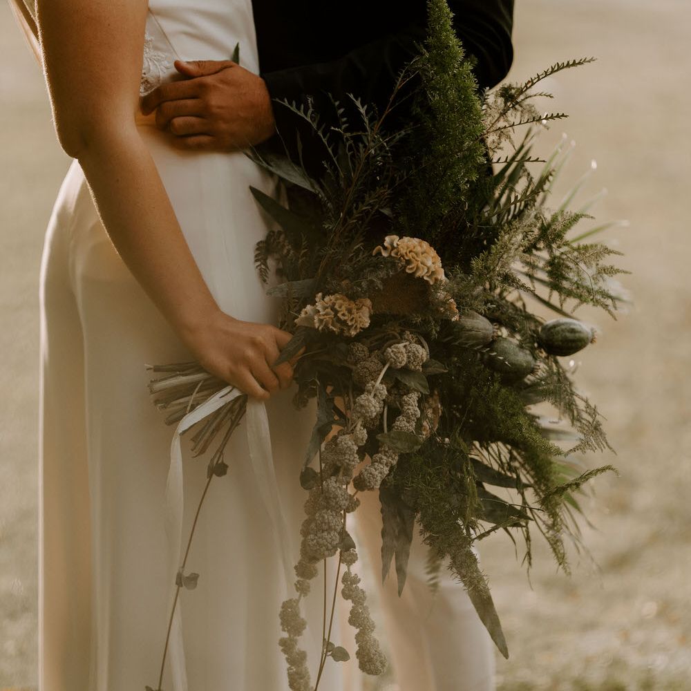 Bride holding a large tropical green wedding bouquet with ferns, dried poppy heads and celosias