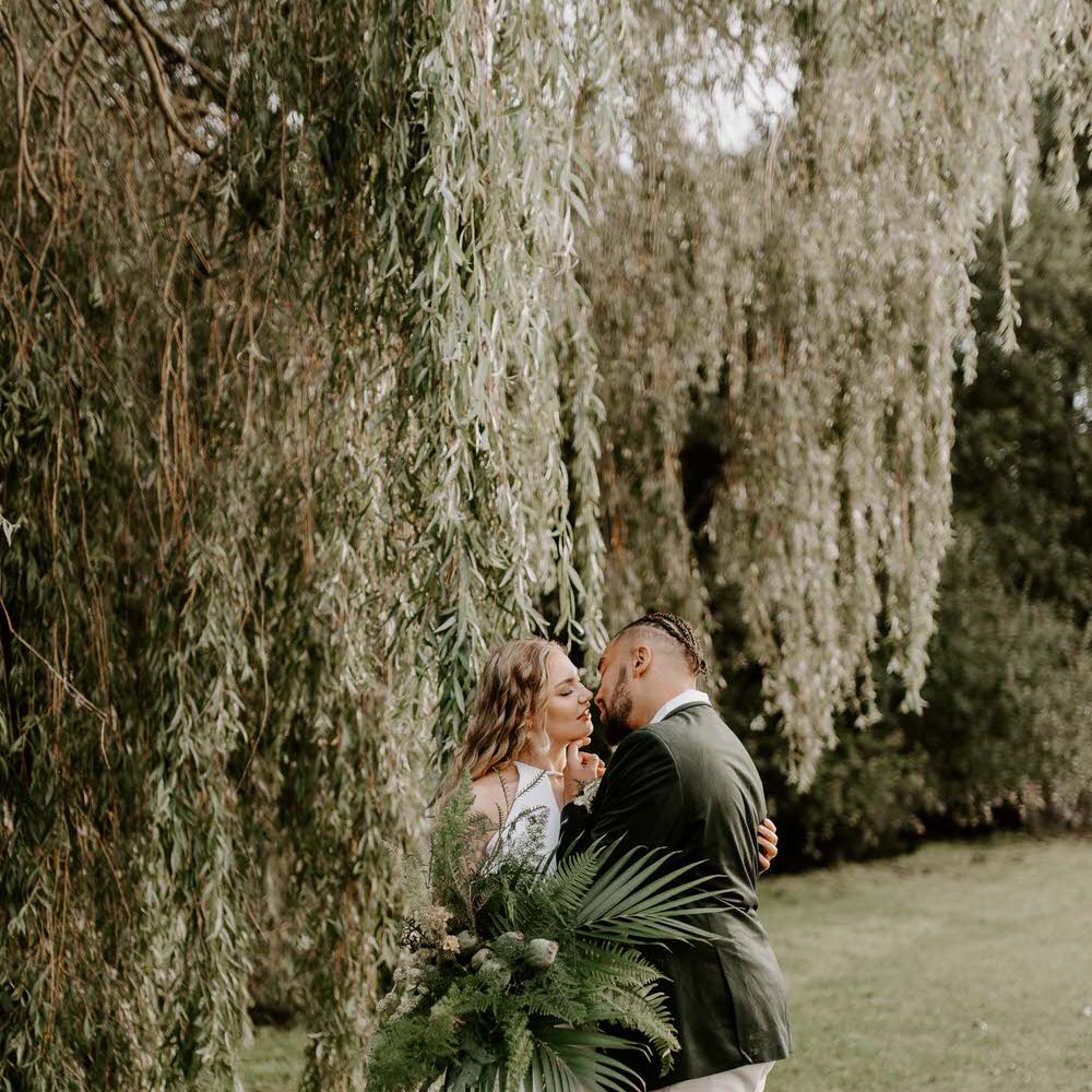 Bride and groom standing under a willow tree, holding a large green tropical bouquet