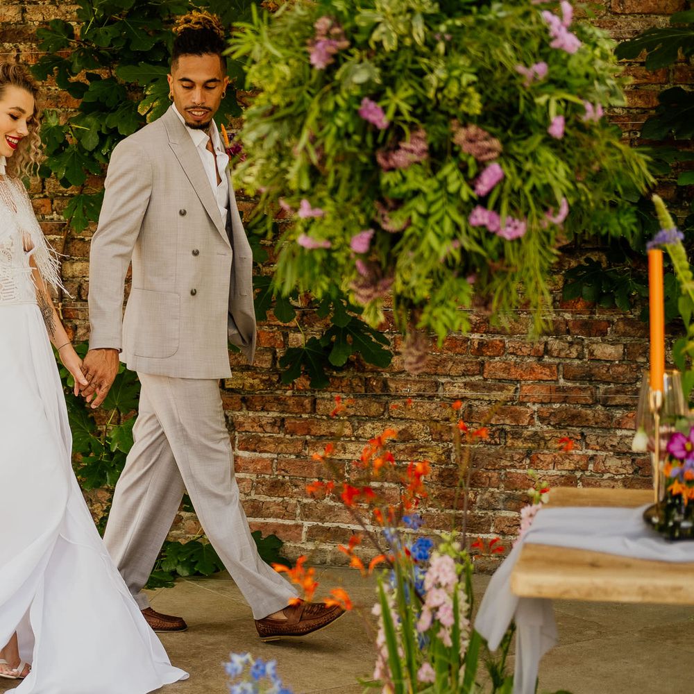 Groom in a beige double breasted jacket holding hands with his bride in bridal separates with tassel coverup