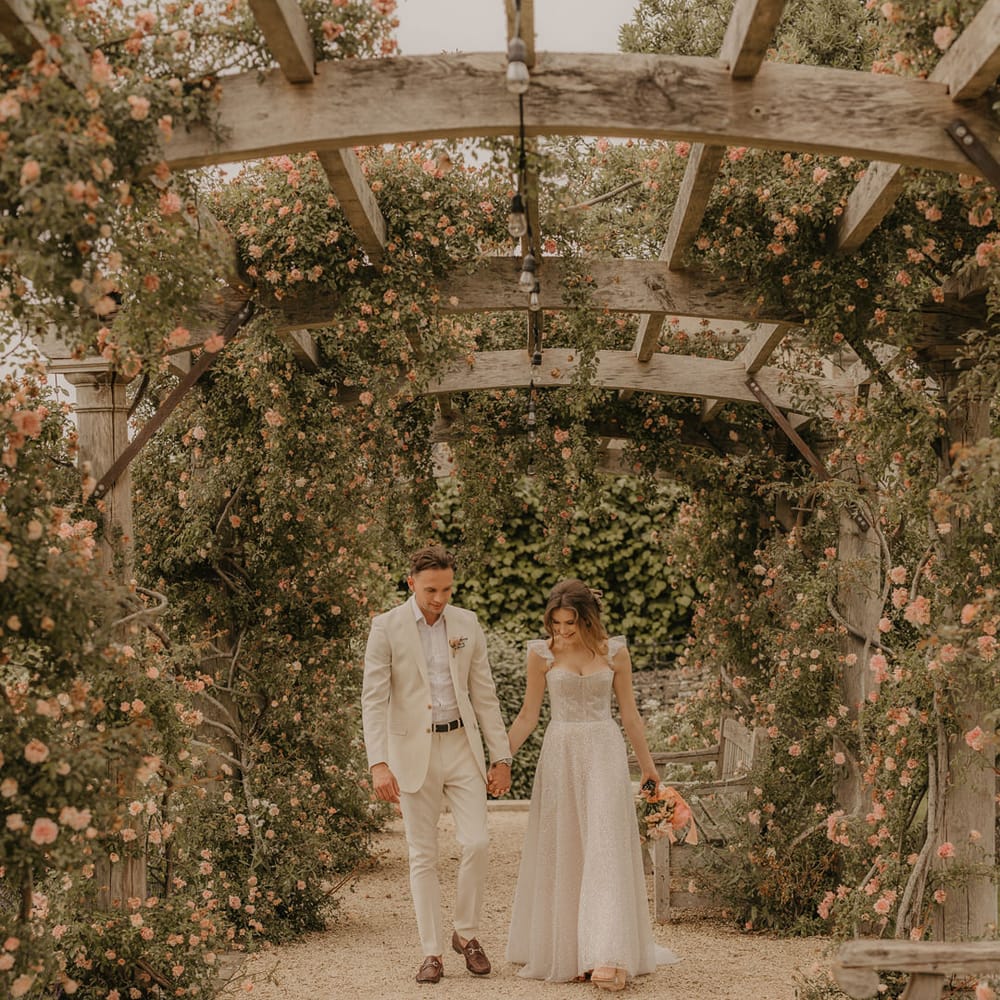 Bride in a sparkly Made With Love dress holding hands with her groom in a stone coloured suit as they walk under the pergola at Euridge Manor 