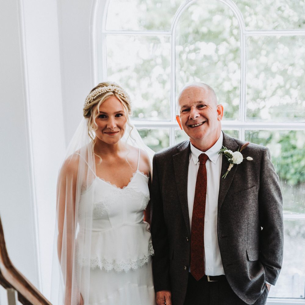 Bride stands with her father on her wedding day in front of window as they prepare to walk down the aisle