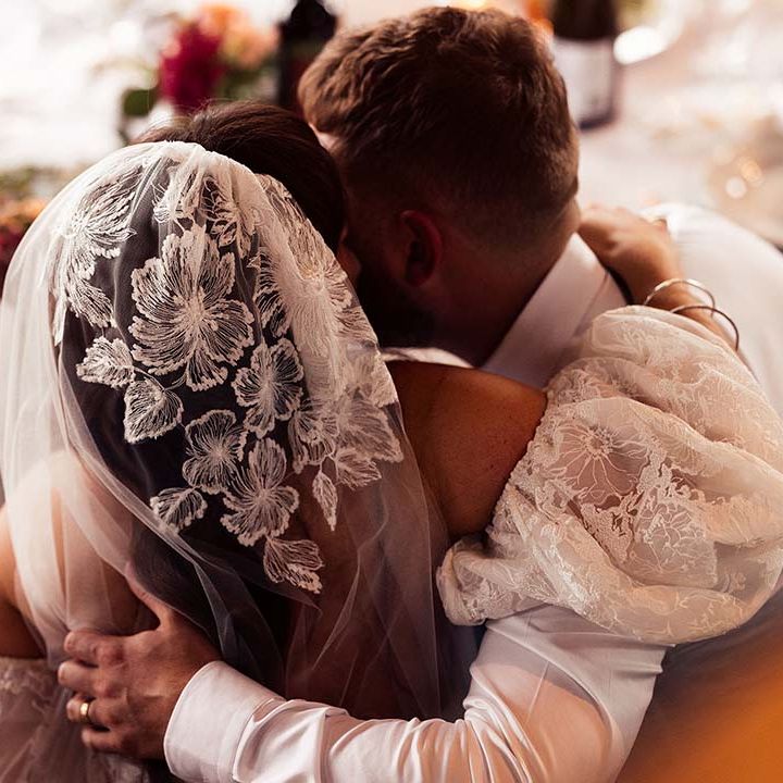 Bride in a lace cap wedding veil with matching detachable puff sleeves embracing her husband at the top table 