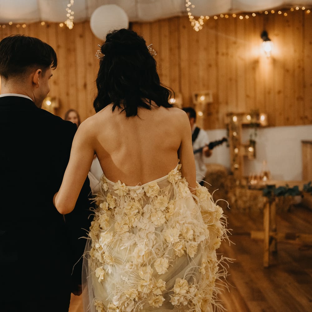 Bride in strapless feathered wedding dress links arm with groom in dark blue suit as they walk down the aisle during rustic wedding ceremony